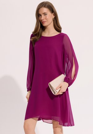 Kobieta w magentowej sukience do kolan z przezroczystymi długimi rękawami, trzyma beżową kopertówkę, stojąc na tle jednolitego jasnego tła.