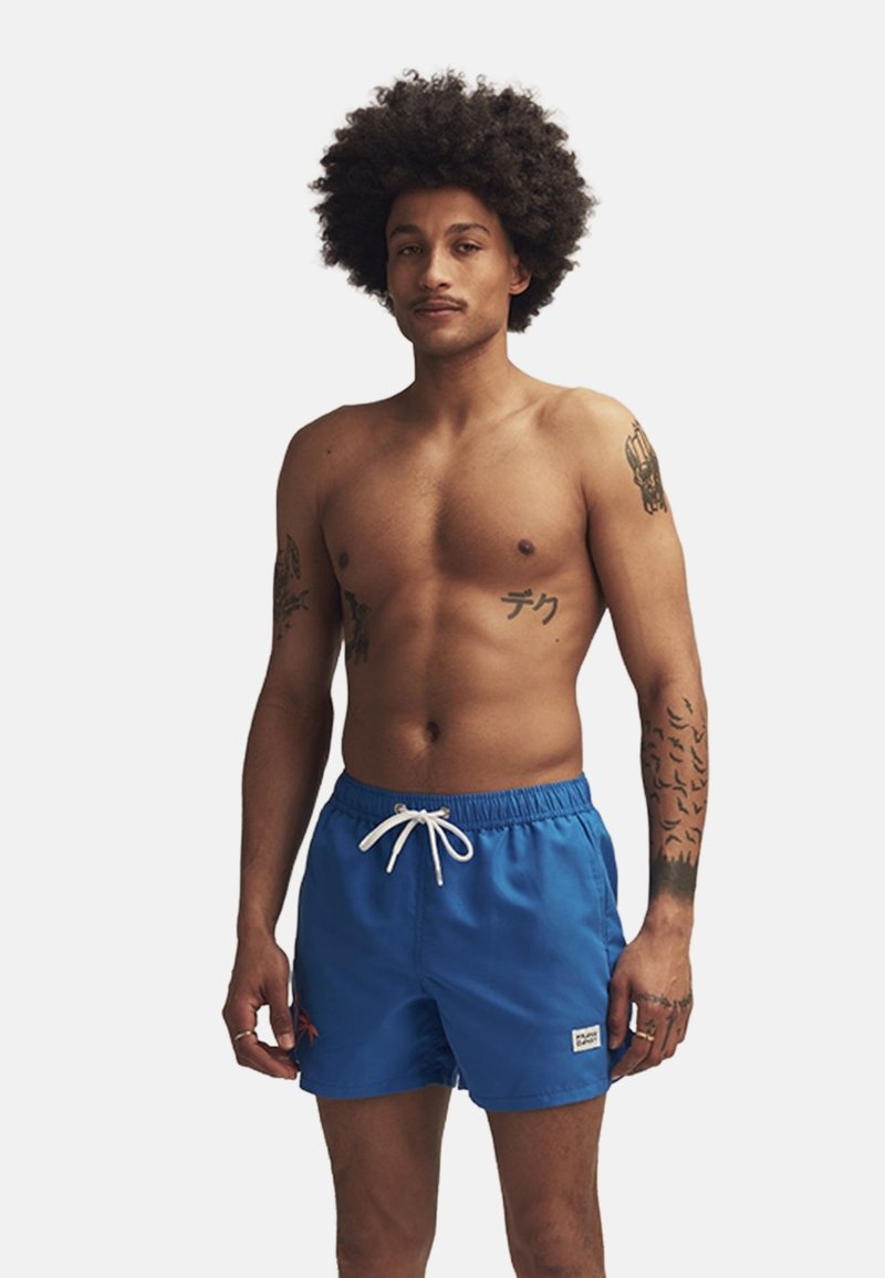 Blå simshorts med elastisk midja, vit dragsko och röd blommig broderi på vänster ben. Modellen är utan tröja.