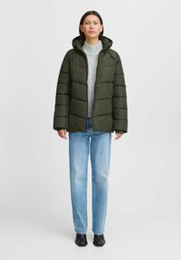 Puhasta jakna v temno zeleni barvi s kapuco, z quiltastim dizajnom in sprednjim zadrgo, nosi čez sivo jopico in svetlo modre jeans.