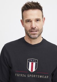 Felpa nera con logo a scudo con "1924" e tre strisce verticali in rosso, bianco e blu; colletto a girocollo a coste e tessuto testurizzato.