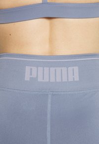 Světle modré sportovní legíny s širokým pasem, na kterém je v jemně růžové barvě logo "PUMA", hladká textura a jemné detaily šití.