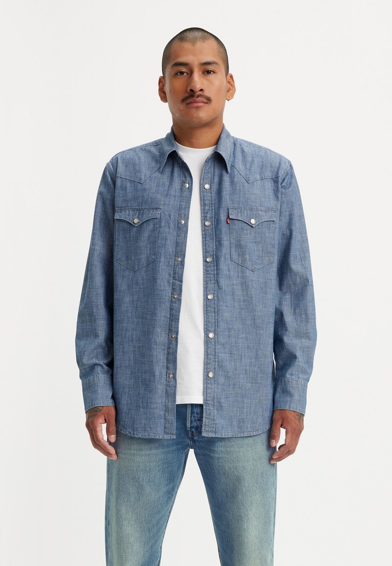 Chemise en denim bleu à boutons, à manches longues, avec des poches poitrine et des accessoires en argent. Portée par-dessus un t-shirt blanc et assortie à un jean clair.