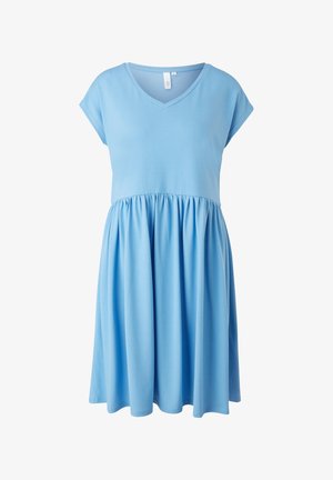 Femme portant une robe bleue mi-longue avec col en V, une veste en jean courte délavée claire et des baskets blanches, debout sur un fond neutre.