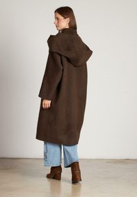 Abrigo de mezcla de lana marrón con capucha grande, corte oversize y mangas anchas, combinado con jeans cortos azul claro y botas de tacón marrones.