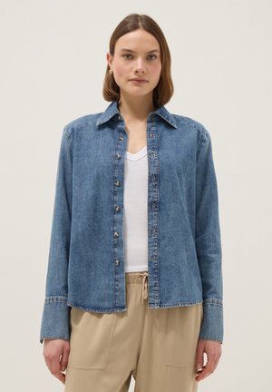 Frau trägt eine offene blaue Jeansjacke über einem weißen V-Ausschnitt-Shirt und beige Hosen mit Kordelzug vor einem einfarbigen Hintergrund.