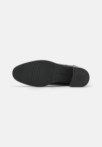 Semelle de chaussure en cuir noir avec une texture lisse, arborant un logo discret et des rainures horizontales pour l'adhérence. Forme courbée et zone de talon renforcée.