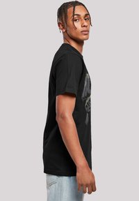 Tricou din bumbac negru cu guler rotund, având un design grafic pe față și mâneci scurte. Modelul poartă blugi de culoare albastră deschisă.