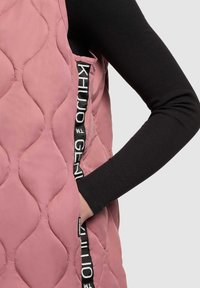 Rosa gepolsterte Weste mit strukturiertem Oberflächen, ausgestattet mit einer Seitentasche und schwarzen Markierungsakzenten am Rand der Tasche. Langer schwarzer Ärmel sichtbar.