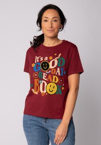 T-shirt en coton bordeaux avec un graphique coloré incluant le texte "C'est une BONNE journée pour LIRE un LIVRE", des visages souriants et des accents floraux.