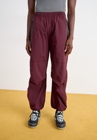 Pantalons légers bordeaux avec une taille élastique, des poches zippées sur les côtés et une coupe décontractée, associés à des chaussures de sport noires et grises.