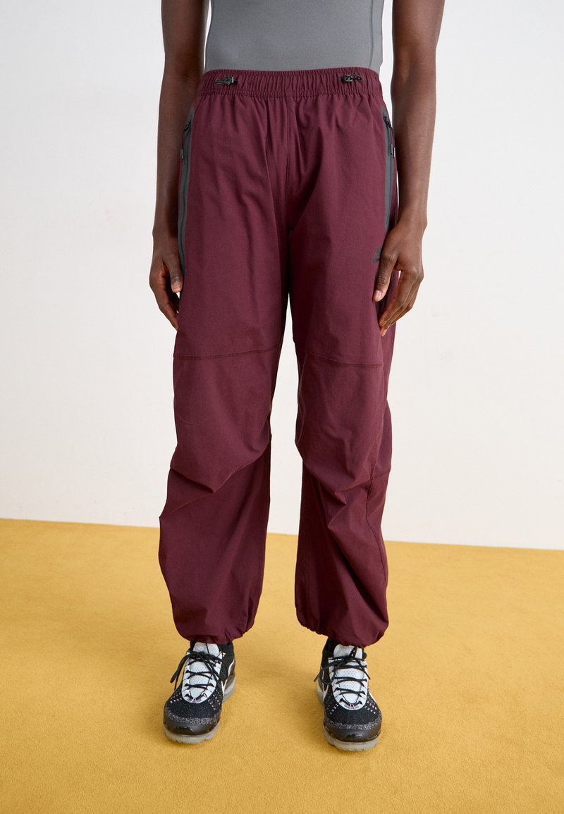 Pantalons légers bordeaux avec une taille élastique, des poches zippées sur les côtés et une coupe décontractée, associés à des chaussures de sport noires et grises.
