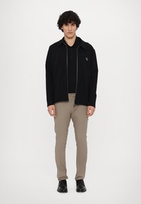 Svart zip-up jacka över en svart polo, tillsammans med beige byxor och mörka skor. Jackan har en subtil logotyp och kontrasterande dragkedja.