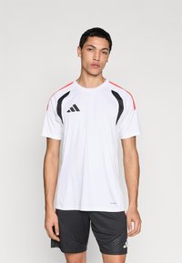 Λευκό αθλητικό t-shirt με μαύρες και κόκκινες λεπτομέρειες, κοντά μανίκια και μαύρο λογότυπο στο στήθος. Φοράται με σκούρο σορτς που έχει σχεδιαστικό μοτίβο.