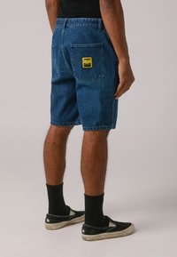 Shorts de mezclilla en azul oscuro con un corte recto, dos bolsillos traseros y una etiqueta amarilla en el bolsillo derecho. Llevados con calcetines y zapatos negros.