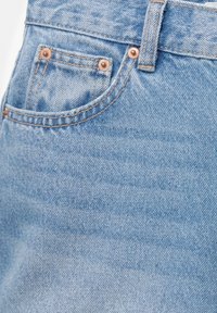 Jean en denim bleu clair en gros plan montrant une poche, une passants de ceinture et des rivets en cuivre sur un tissu texturé.