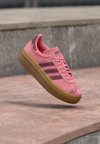 Ροζ αθλητικό παπούτσι Adidas Gazelle με καφέ σόλα και ρίγες, που αιωρείται πάνω από πέτρινα σκαλοπάτια.
