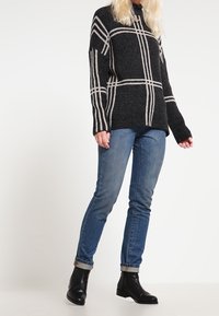 Ciemnoszary sweter w biały wzór w kratę, zestawiony z niebieskimi dżinsami i czarnymi botkami za kostkę. Sweter ma luźny krój i długie rękawy.