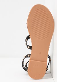 Sandale plate noire avec clous dorés, montrant une semelle et un talon texturés couleur tan, taille 4.