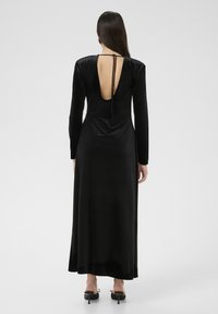 Robe en velours noir à manches longues, avec un décolleté profond dans le dos, longueur cheville. Présente une fermeture nouée dans le dos. Texture lisse.