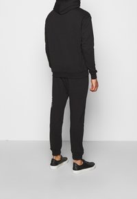 Sudadera negra de algodón con un bolsillo tipo canguro y puños de canalé, combinada con pantalones jogging a juego que cuentan con una cinturilla elástica. Se lleva con zapatillas negras.
