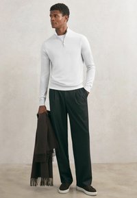 Pulover tricotat alb cu guler cu fermoar, pantaloni negri lejeri, ținând o eșarfă maro închis cu franjuri, asortată cu adidași închiși la culoare, cu textură netedă în ansamblu.