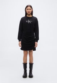 Μαύρο φόρεμα με μακριά μανίκια και ζώνη στη μέση, με γκρι λογότυπο Calvin Klein. Φορεμένο με μαύρες χοντρές μπότες, λείο ύφασμα.