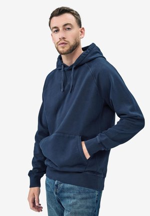 Uomo con capelli corti castani e barba, indossa una felpa con cappuccio blu navy e jeans blu, posa con una mano in tasca su sfondo bianco.