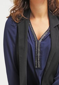 Blusa blu navy con scollo a V profondo e ricami argentati, abbinata a un blazer nero. Tessuto morbido, vestibilità rilassata e dettagli eleganti.