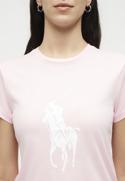 Γυναίκα που φοράει ανοιχτό ροζ μπλουζάκι με λευκό γραφικό παίκτη πόλο που καβαλάει άλογο και κουνάει mallet, εμφανίζεται από το λαιμό έως το μέσο του κορμού.