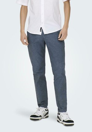 Pantaloni slim-fit blu scuro con una texture liscia, caratterizzati da un sottile motivo a intreccio. Abbinati a sneaker bianche e una camicia bianca con bottoni.