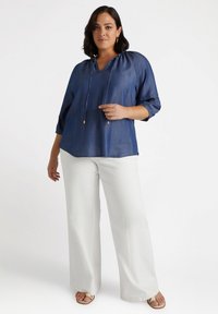 Pullover blu denim con scollo a V, cappuccio con cordino e maniche 3/4; abbinato a pantaloni bianchi a gamba larga e sandali.