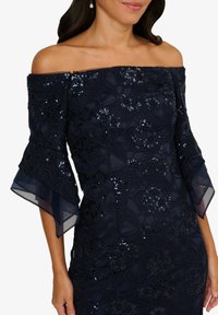 Abito navy off-the-shoulder con ricamo in pizzo floreale, paillettes e maniche a campana trasparenti. La texture del tessuto liscio esalta il design elegante.