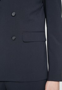Mornariški krojen blazer z dvojimi gumbi, enim žepom in strukturiranim dizajnom iz gladkega, lahkega materiala.