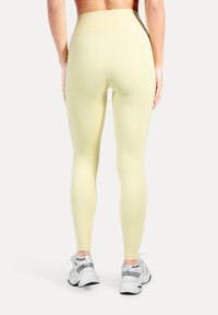 Ljusgula, höga, midjade leggings med en slät, elastisk textur. Bärs med vita sportskor, bakifrån.