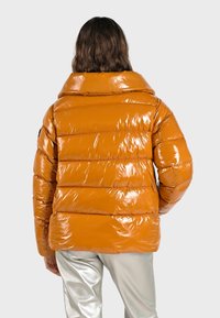 Doudoune orange avec une finition brillante, dotée d'un col montant et d'un design matelassé horizontal. Portée avec un pantalon argent métallique.