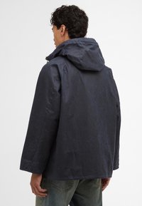 Casaco impermeável azul-marinho com capuz, apresentando um acabamento texturizado, corte solto e punhos com botão. A vista traseira mostra um design minimalista.