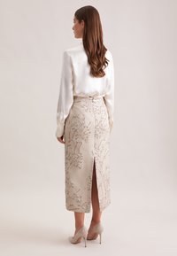 Donna con lunghi capelli castani, vestita con una blusa di satin bianca e una gonna midi beige ricamata a fiori con spacco posteriore e tacchi alti beige.