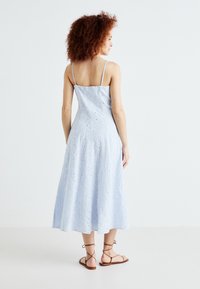 Rochie midi bleu deschis cu bretele subțiri, țesătură texturată cu accente brodate și tiv evazat. Asortată cu sandale maro cu curele.