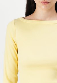 Sárga bordás top, csónaknyakkal, hosszú ujjakkal és sima textúrával. Ez a ruhadarab fekvő siluettet biztosít.