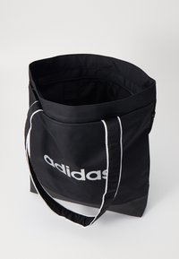 Black nylon tote bag with white piping and "adidas" logo. Open top, rectangular shape, dual shoulder straps, and smooth texture.
