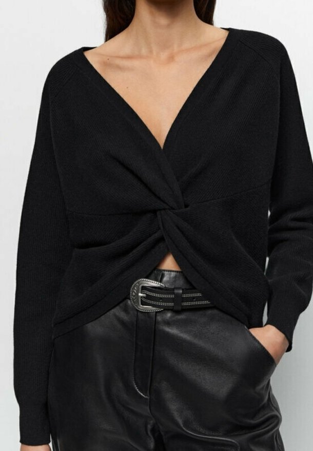 Femme portant un pull noir à manches longues avec un col en V torsadé sur le devant et un pantalon en cuir noir avec une ceinture, main dans la poche, torse visible.
