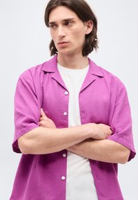 Νέος άνδρας με καστανά μαλλιά μέχρι τον ώμο, φοράει λευκό μπλουζάκι κάτω από μωβ κοντομάνικο πουκάμισο με κουμπιά, σταυρωμένα τα χέρια, κοιτάει αριστερά.