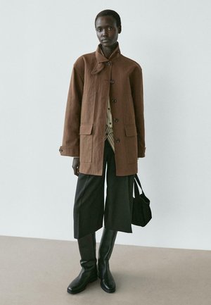 Persona alta che indossa un cappotto marrone con bottoni sopra una camicia a righe, pantaloni neri larghi, stivali neri fino al ginocchio, tiene una borsa nera, in piedi contro uno sfondo semplice.