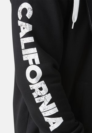 Sweat à capuche noir avec l'inscription "CALIFORNIA" en blanc imprimée dans un style usé le long de la manche gauche. Comprend une fermeture éclair à l'avant et une capuche avec cordon.