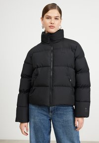 Giubbotto piumino nero con colletto alto, chiusura con zip e due tasche laterali, caratterizzato da un motivo a trapunte e logo Lacoste sul petto.