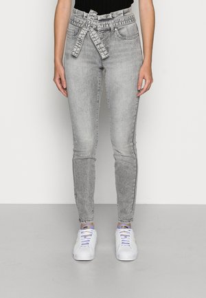 Henkilö pukeutuneena vaaleanharmaisiin korkeavyötäröisiin farkkuihin, joissa on solmittu vyö, ja valkoisiin tenniskengiin, seisoo yksinkertaisen vaalean taustan edessä.