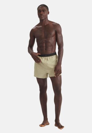 Athletischer dunkelhäutiger Mann steht barfuß, trägt beige Shorts mit schwarzem Bund und posiert mit einer Hand nahe dem Bund vor einfachem Hintergrund.