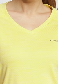 Żółta koszulka z dekoltem w serek o teksturowanym wzorze, z małym logo Columbia nadrukowanym po lewej stronie klatki piersiowej. Lekki, casualowy design.