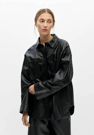 Femme aux cheveux attachés portant une chemise et un pantalon en cuir noir oversize, regardant sur le côté sur un fond blanc uni.