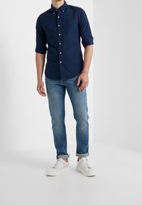 Námořnická košile s knoflíky a ohrnutými rukávy, světle modré džíny a bílé tenisky. Košile má na hrudi malé logo.